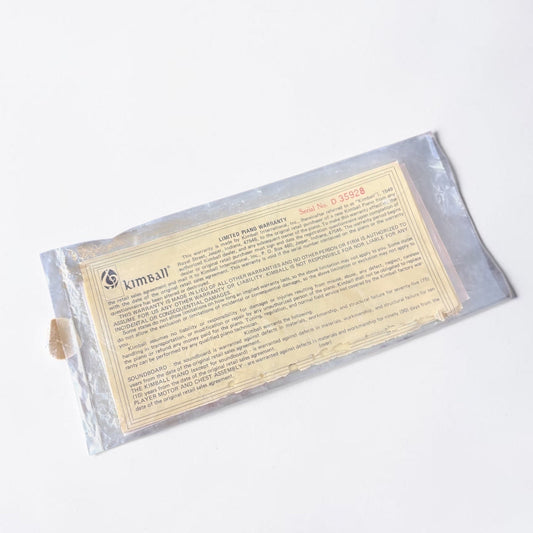 Outrageous Art vintage Kimball Piano limited warranty document in a protective plastic sleeve on a wooden surface.